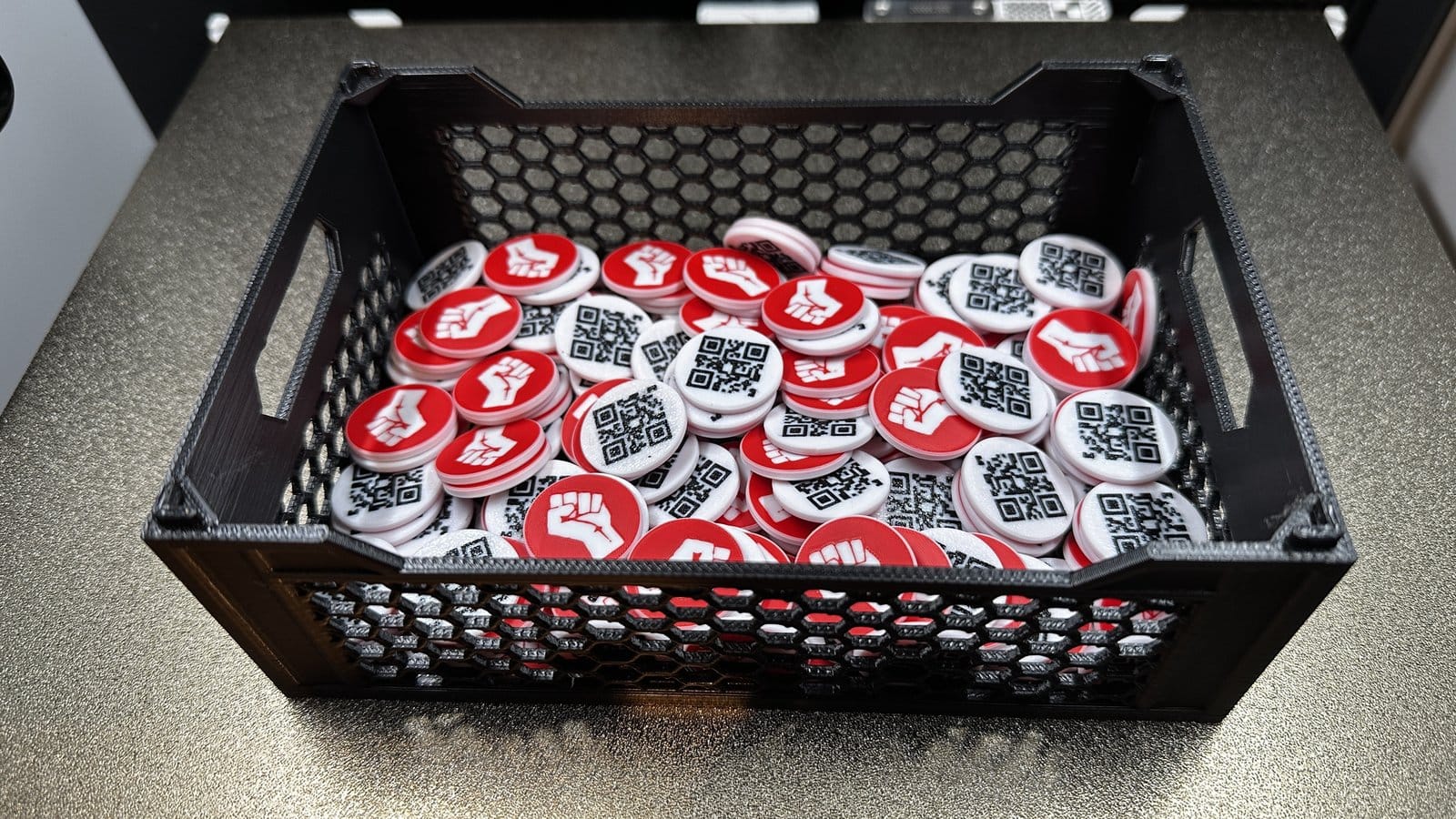 Transportkiste voller 3D-gedruckter Einkaufswagenchips mit erhobener Faust und QR-Code auf der Rückseite